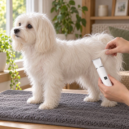 White fluffy dog standing on a grey mat while hands hold the trimmer against its coat