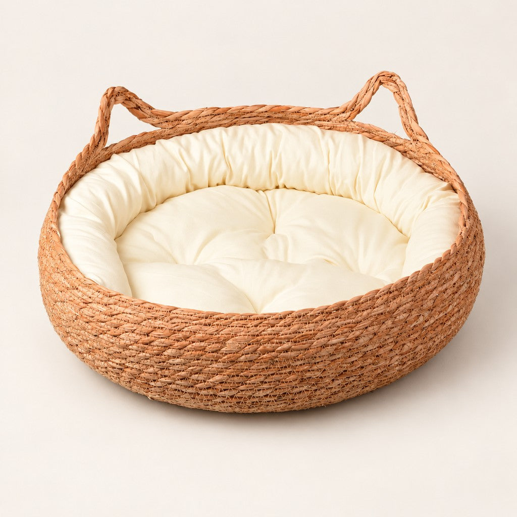 Round woven straw cat basket in orange-brown with cream cushion and two looped handles on pale background