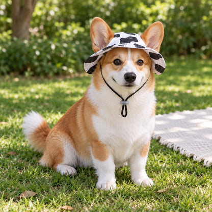 Corgi sitting on the lawn beside a cream blanket, wearing the cow-print bucket hat